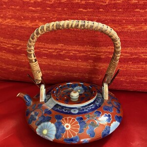Puede incluir: Una tetera de cerámica con asa trenzada. La tetera presenta una base roja con diseños florales azules y naranjas. La tapa tiene un patrón a juego y un pequeño pomo blanco. El asa es de ratán trenzado.