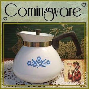 Puede incluir: Una tetera CorningWare blanca con un patrón floral azul en la parte frontal, una banda metálica alrededor del cuello, un asa marrón oscuro y una tapa a juego con un pequeño pomo marrón. La palabra CorningWare está impresa en negro arriba.