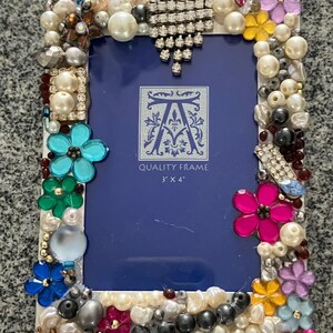 May include: A white picture frame decorated with pearls, rhinestones, and colorful flowers. The frame is 3 inches by 4 inches and says "Quality Frame" on the front.