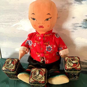 May include: A vintage doll with a painted face, wearing a red jacket with floral patterns and black pants. The doll has a topknot hairstyle and is seated next to three decorative black and red tins.
