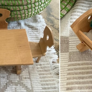 Może przedstawiać: Drewniany zestaw mebli do domku dla lalek z stołem i dwoma krzesłami oraz kołyską z lalką w środku. Lalka ma na sobie zielony kapelusz i białą koszulę z czarnym kamizelką.