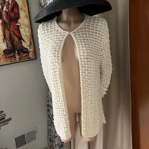 Puede incluir: Un cárdigan crema con frente abierto y un diseño de tejido abierto texturizado. El cárdigan se exhibe en un maniquí, combinado con un sombrero negro de ala ancha adornado con perlas y plumas. El sombrero es un accesorio elegante.