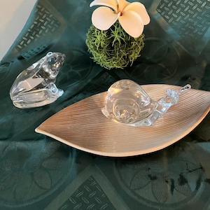 Puede incluir: Una figura de rana de cristal transparente y una figura de caracol de cristal sobre una bandeja con forma de hoja. Una bola de musgo con una flor crema se encuentra en el fondo. Sobre un mantel verde oscuro.
