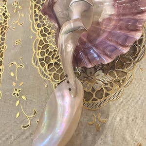 Pode incluir: Objeto decorativo em forma de concha com acabamento perolado. O objeto apresenta uma grande concha alongada e iridescente, presa a uma base em forma de concha com um esquema de cores rosa e branco. O design inclui uma forma curva e elegante.