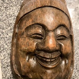 Könnte beinhalten: Eine geschnitzte Holzmaske, die ein lächelndes Gesicht mit Hut darstellt. Die braune Maske hat sichtbare Holzmaserung und einen fröhlichen Ausdruck. Die Maske hat zwei kleine Knöpfe auf der Hutoberseite.