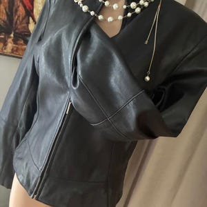 Peut inclure: Une veste en cuir noir avec un col montant et une fermeture éclair frontale intégrale. La veste est accessoirisée avec un collier de perles blanches superposées, un pendentif floral doré et une longue chaîne fine avec une perle unique.