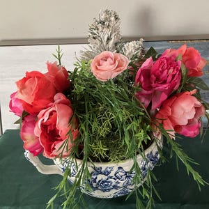 Puede incluir: Un arreglo floral con vibrantes peonías rosas y rojas, una rosa rosa pálido y follaje verde. Las flores están dispuestas en una tetera con estampado azul y blanco, creando un centro de mesa encantador.