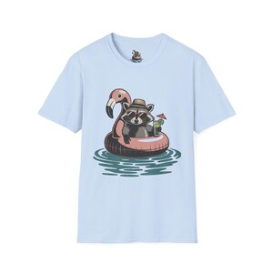 Peut inclure: T-shirt bleu clair avec un raton laveur de dessin animé portant des lunettes de soleil et un chapeau, flottant sur un bouée gonflable flamant rose. Le raton laveur tient un cocktail avec un petit parapluie. Le motif est sur un fond d'eau.