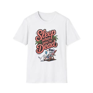 May include: White t-shirt featuring a skeleton relaxing in a beach chair, wearing sunglasses and holding a drink. The shirt has the text "Sleep when you're Dead" in a stylized font with palm trees.