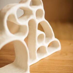 May include: A white ceramic sculpture with a porous, organic structure featuring multiple interconnected arched openings and a speckled matte finish. The object has a curved, layered design that creates a series of hollow voids throughout its form.