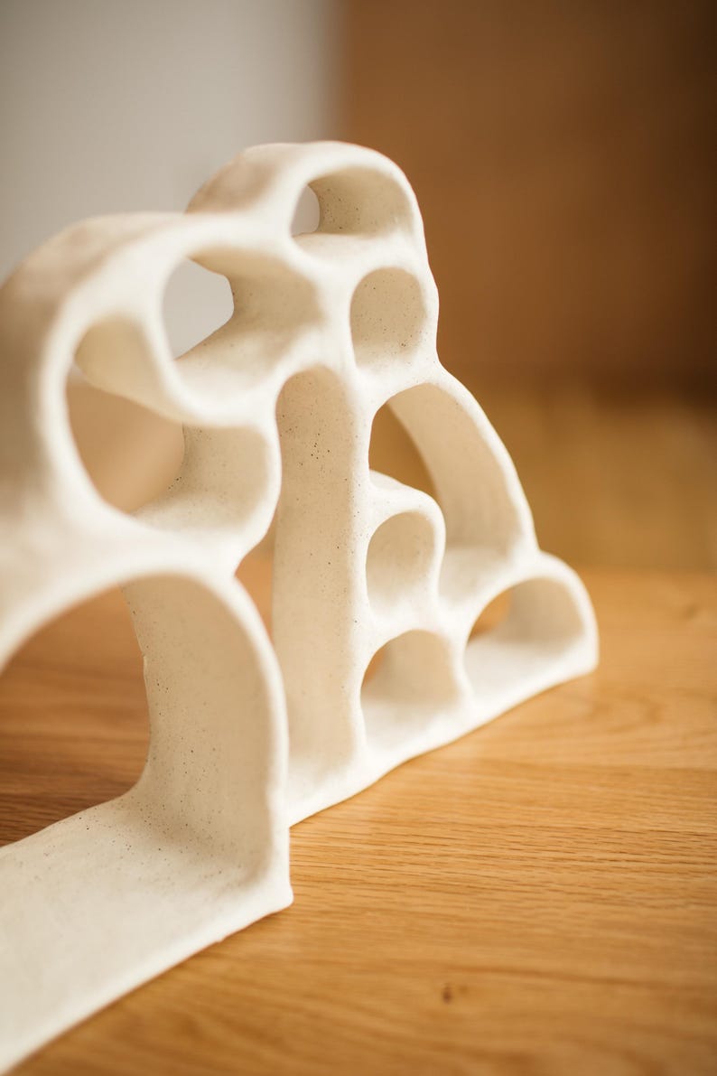 May include: A white ceramic sculpture with a porous, organic structure featuring multiple interconnected arched openings and a speckled matte finish. The object has a curved, layered design that creates a series of hollow voids throughout its form.