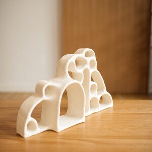 May include: A white ceramic sculpture featuring an organic, multi-arched structure with various circular and semi-circular cutouts. The piece has a matte, textured finish and a series of interconnected openings that create a hollow, layered form.