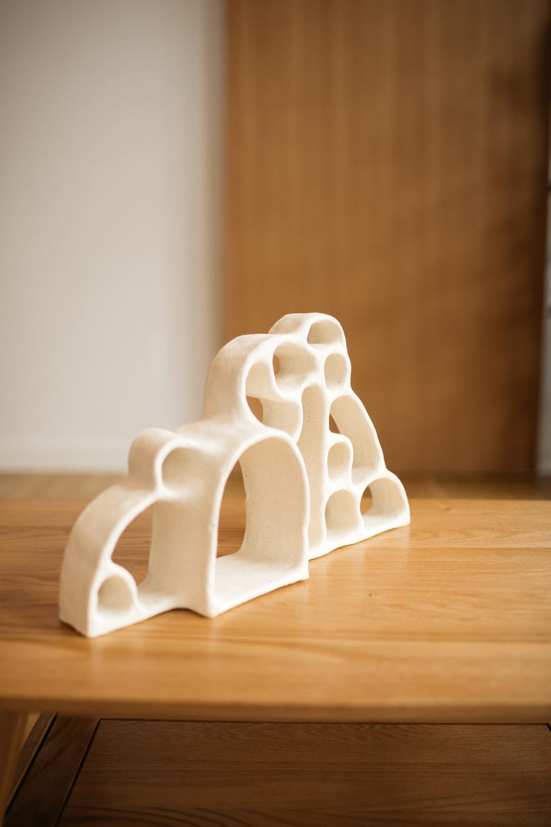 May include: A white ceramic sculpture featuring an organic, multi-arched structure with various circular and semi-circular cutouts. The piece has a matte, textured finish and a series of interconnected openings that create a hollow, layered form.