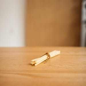 May include: A bundle of light-colored reed diffuser sticks tied together with twine, resting on a light-colored wooden surface. The background features a blurred neutral color palette.