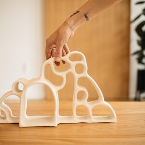 May include: A white ceramic sculpture featuring an organic, open-work design with multiple rounded cutouts and a smooth, matte finish. The structure has a wide, arched base and an irregular, flowing silhouette that creates a series of interconnected loops.