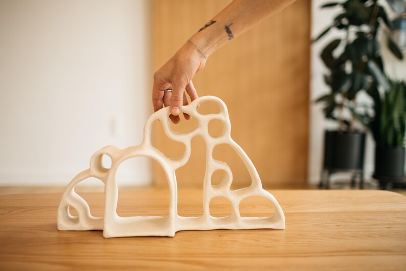 May include: A white ceramic sculpture featuring an organic, open-work design with multiple rounded cutouts and a smooth, matte finish. The structure has a wide, arched base and an irregular, flowing silhouette that creates a series of interconnected loops.