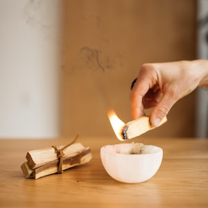 May include: A lit palo santo stick held over a white ceramic bowl, with smoke rising. A bundle of palo santo sticks tied with twine sits nearby on a wooden surface. The flame is bright orange.