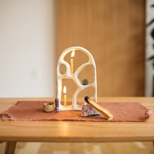 May include: A cream-colored, abstract candle holder with three lit candles. The holder sits on a terracotta-colored runner on a wooden table, alongside crystals, a small woven basket, and a piece of Palo Santo.