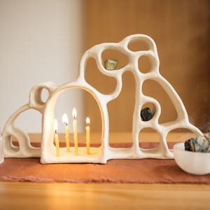 May include: A cream-colored, organic-shaped ceramic candle holder with multiple arched openings, holding four lit thin yellow candles. A white crystal tower, a small white bowl with a sage bundle, and two small stones are placed nearby.