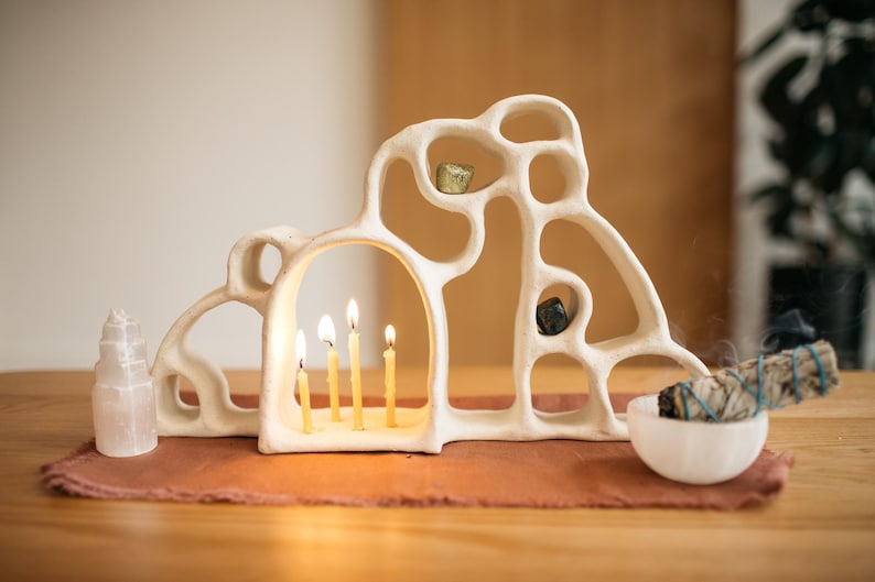 May include: A cream-colored, organic-shaped ceramic candle holder with multiple arched openings, holding four lit thin yellow candles. A white crystal tower, a small white bowl with a sage bundle, and two small stones are placed nearby.