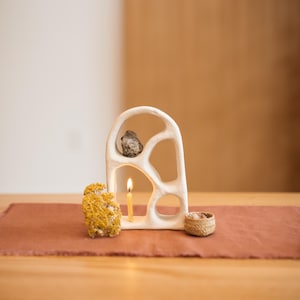 May include: A white ceramic sculpture with organic cutouts, holding a small rock and a lit candle. A small woven basket and dried yellow flowers are on a rust-colored runner. The scene is set on a wooden table.