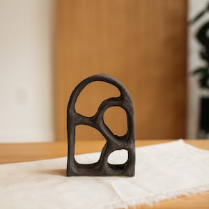 May include: A dark brown, abstract sculpture with an arched top and several irregular openings. The art piece is made of a textured material and sits on a white cloth, with a wooden background.
