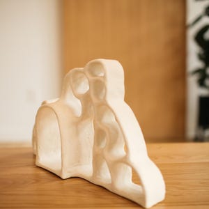 May include: A cream-colored ceramic sculpture featuring an organic, porous structure with multiple rounded openings and a smooth, matte finish. The object has a curved, asymmetrical silhouette with interconnected hollow sections throughout its form.