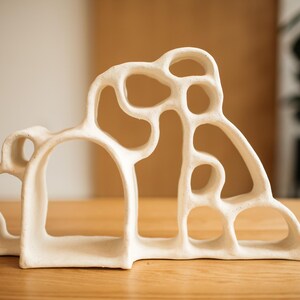 May include: A cream-colored ceramic sculpture featuring an organic, open-work design with multiple interconnected rounded arches and varying circular voids. The structure has a matte, textured surface and a wide, stable base with a continuous form.