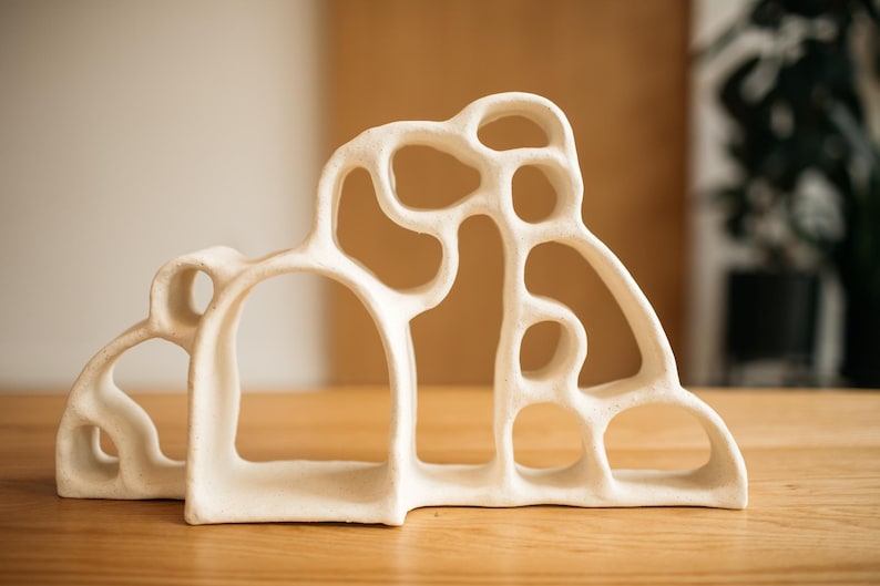 May include: A cream-colored ceramic sculpture featuring an organic, open-work design with multiple interconnected rounded arches and varying circular voids. The structure has a matte, textured surface and a wide, stable base with a continuous form.