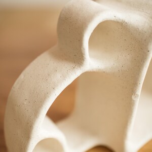 May include: A sculptural off-white ceramic object with a matte, speckled finish. The piece features organic, curved forms and hollow, arched openings that create a fluid, interconnected structure with a textured, porous surface appearance.