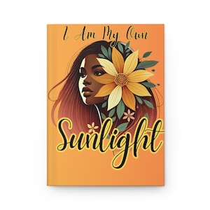 Puede incluir: Un diario de tapa dura con una cubierta naranja que presenta una ilustración de una mujer con cabello castaño largo y una gran flor amarilla. El texto "I Am My Own Sunlight" está escrito en una fuente de guion.