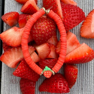 May include: A red and orange beaded bracelet with a small strawberry charm. The bracelet is surrounded by sliced strawberries.