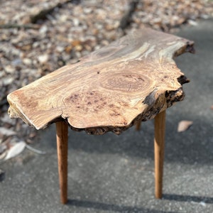 May include: A rustic wooden side table with a live edge top and two wooden legs. The table is made of natural wood with a unique grain pattern.