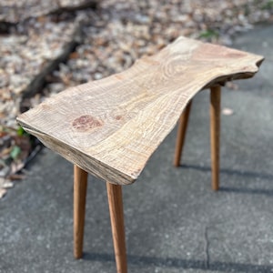 Puede incluir: Una mesa auxiliar de madera rústica con una tapa de borde vivo y cuatro patas delgadas. La mesa está hecha de madera marrón claro y tiene un aspecto natural y sin terminar.