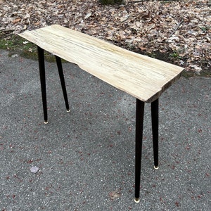 Puede incluir: Una mesa consola de madera rústica con una tapa de borde vivo y patas de horquilla negras con puntas doradas.