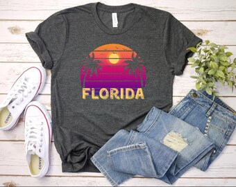 Puede incluir: Una camiseta gris oscuro con un gráfico de puesta de sol retro y palmeras. El texto "FLORIDA" está impreso en amarillo en la camiseta.