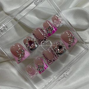 May include: A set of pink and silver press-on nails with rhinestones and star charms. The nails are displayed in a clear plastic case.