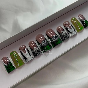 May include: A set of ten green and white press-on nails with silver accents. The nails feature a variety of designs, including a cow print, a cross, and a planet. The nails are displayed in a white box.