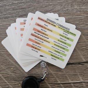 May include: A stack of white cards with Spanish commands printed on them, attached to a black retractable badge reel. The cards have phrases like "Please" and "Thank you" in English and Spanish.