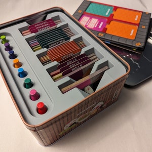 May include: A metal game box with a bamboo design and a sushi theme. The box contains cards with different categories, including "Special Rolls", "Rolls", "Appetizer", "Dessert", and "Recent". The box also includes a set of colorful wooden markers.
