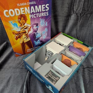 May include: Codenames Pictures board game box and open game tray with organized card slots. The box cover features the title Codenames Pictures, the author Vlaada Chvátil, and illustrations of two figures interacting with holographic blue icons.