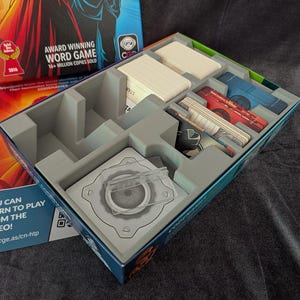 May include: The Codenames board game box features a gray plastic insert with organized compartments for game components. It includes stacks of white cards, blue cards, red cards, and a central square tile holder with a clear plastic stand.
