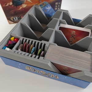 May include: A gray and blue organizer for the Dixit board game. The organizer has multiple compartments for cards, tokens, and other game components. The Dixit logo is visible on the side of the organizer. The game box is visible in the background.