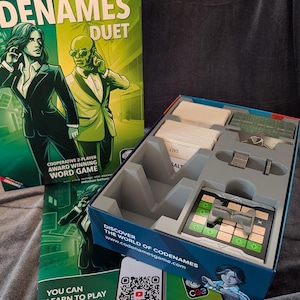 May include: Codenames Duet board game box and open game tray. The box features green illustrations of two figures in suits. The open tray contains stacks of white word cards, a green card deck, a grid board with a gray plastic stand, and text.