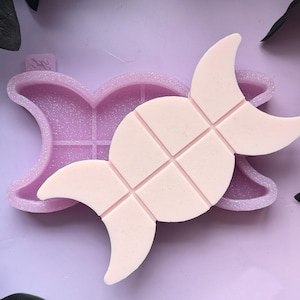 May include: A pink silicone mold shaped like a triple moon with a grid pattern. The mold is filled with a light pink wax.