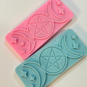 May include: Two rectangular, decorative soap bars. One is pink, the other is blue. Each bar features a pentagram within a circle, flanked by crescent moons and stars. The bars are likely handmade and suitable for spiritual or Wiccan practices.