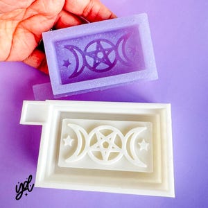 May include: Two silicone molds for resin casting. The top mold is purple and the bottom mold is white. Both molds feature a triple moon and pentagram design.