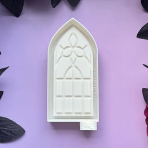 May include: White plastic mold of a gothic arch window with a decorative design. The mold is approximately 10 inches tall and 6 inches wide.