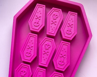 May include: A pink silicone mold shaped like a coffin with nine individual molds inside, each shaped like a coffin with a skull and crossbones design.