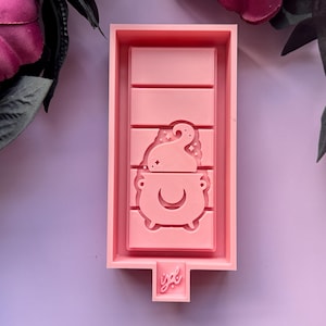 May include: Pink silicone mold for resin casting, featuring a cauldron with a swirling steam design. The mold has a rectangular shape with a raised section for the cauldron design. The mold is labeled "ygl".